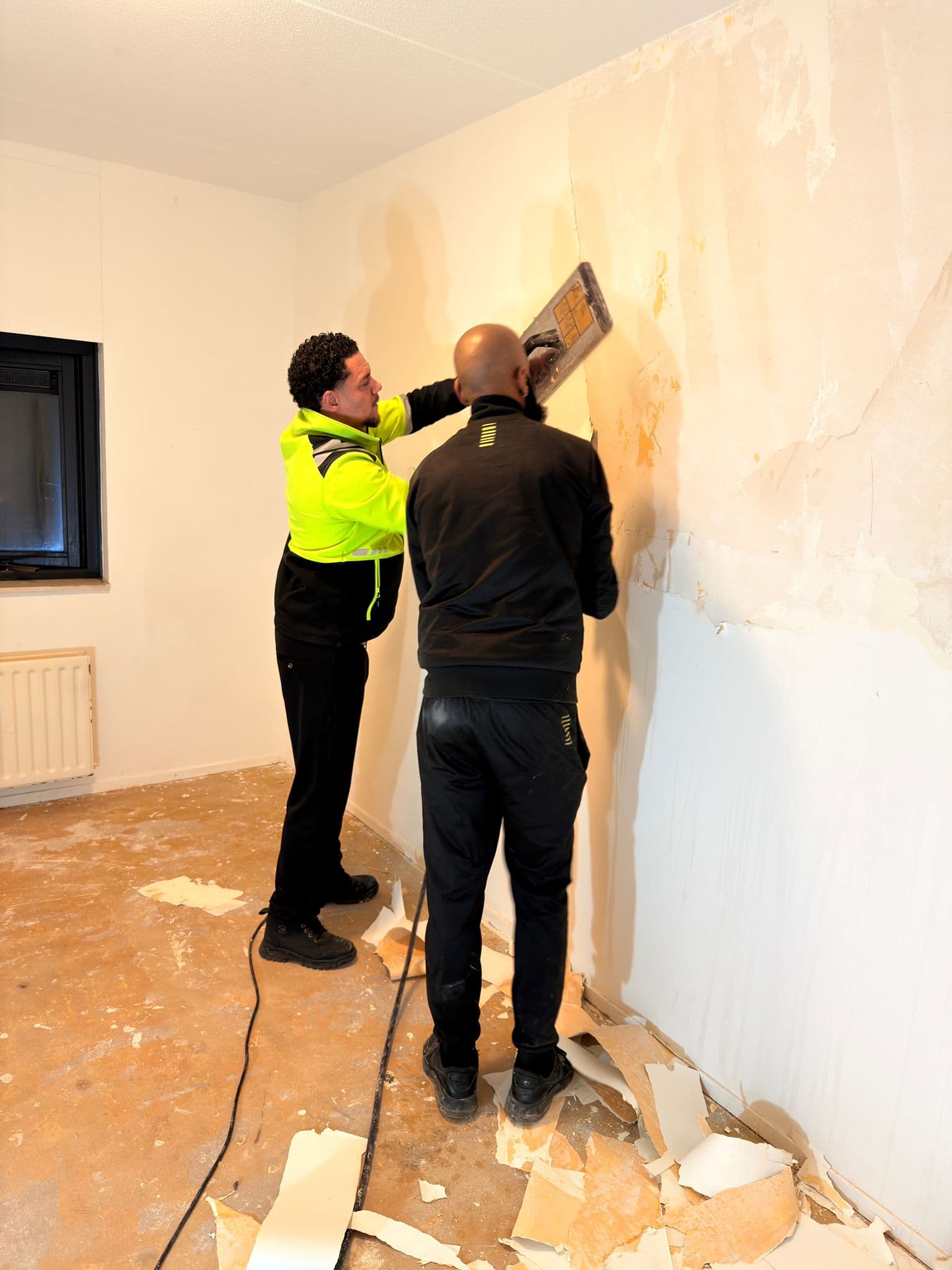 Two men stripping old wallpaper from a wall in a room undergoing renovation.