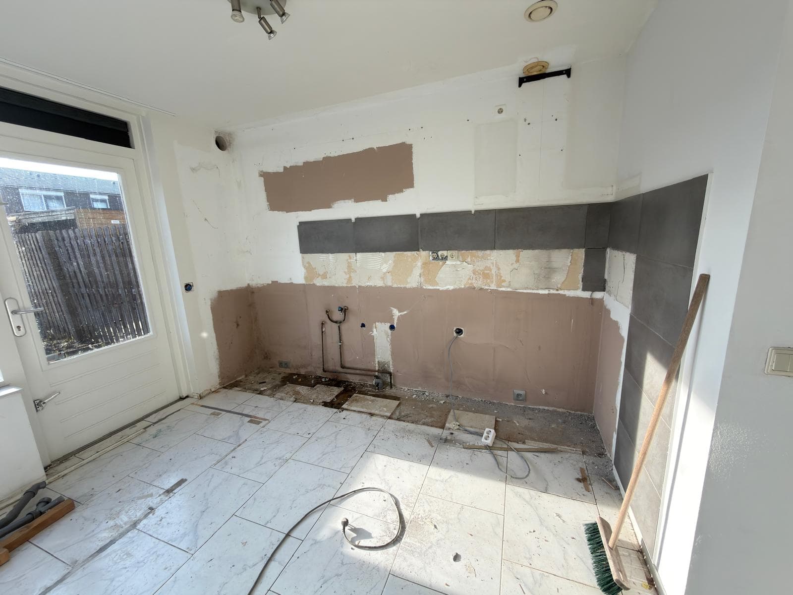 A kitchen undergoing renovation with stripped walls, exposed pipes, and white marble-look floor tiles.