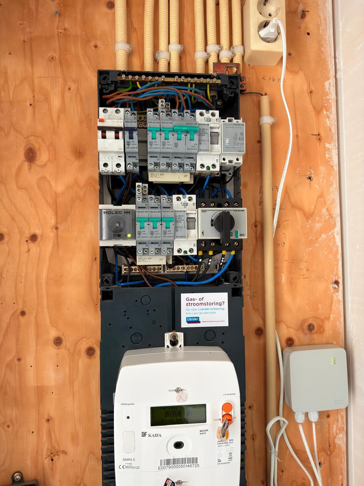 Electrical panel with circuit breakers, exposed wiring, and smart meter on a plywood wall.